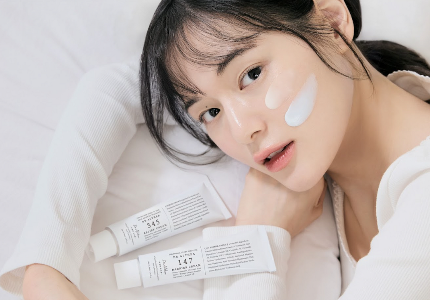 Meitene guļ uz balta fona ar krēma paraugiem uz vaiga, blakus redzamas ādas kopšanas krēmu tūbiņas Dr. Althea 147 un 345.