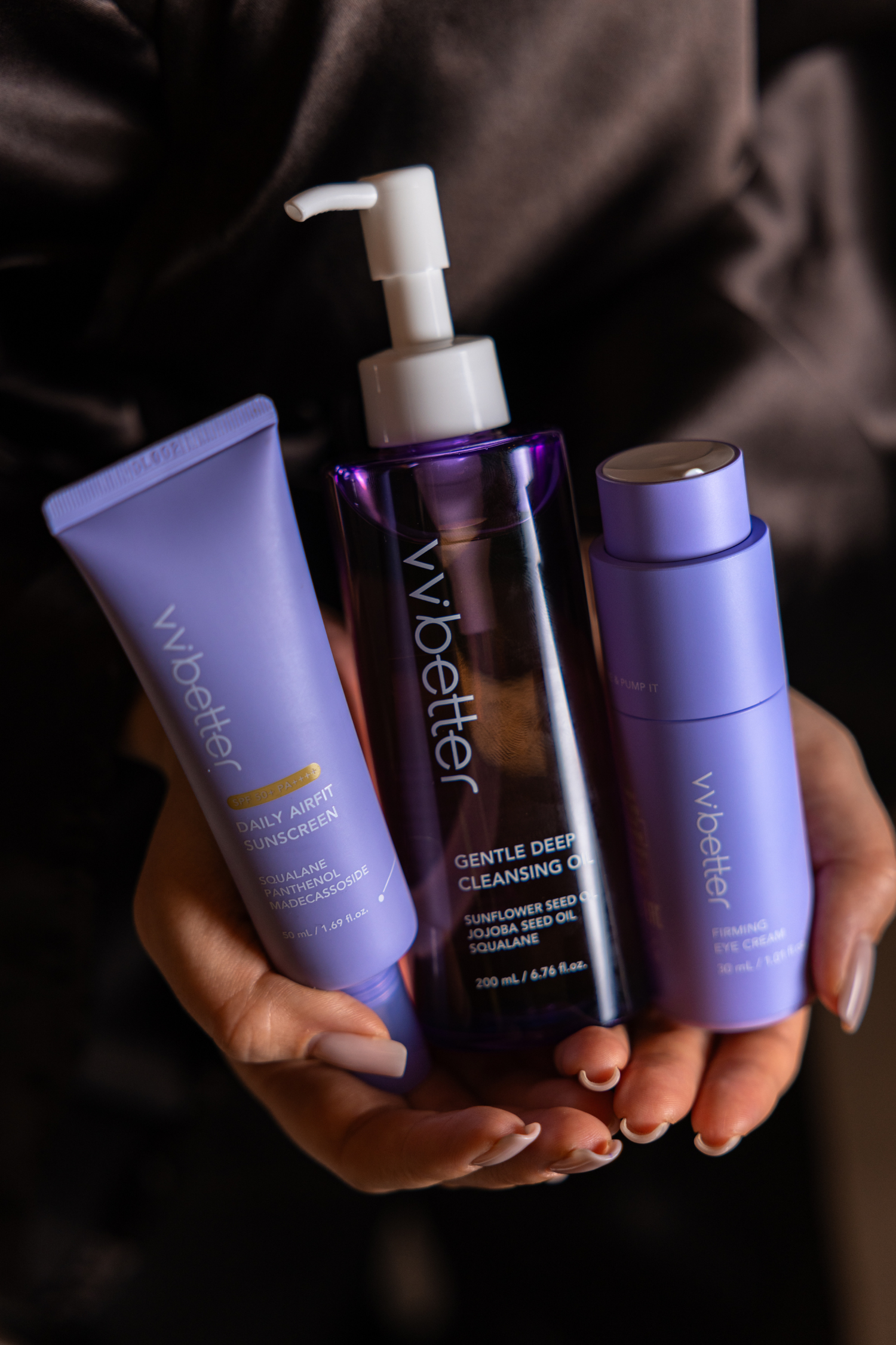 Rokās turēti trīs VVBetter produkti violetā iepakojumā, saules aizsargkrēms, attīrošā eļļa ar pumpīti un acu krēms, tumšs fons.